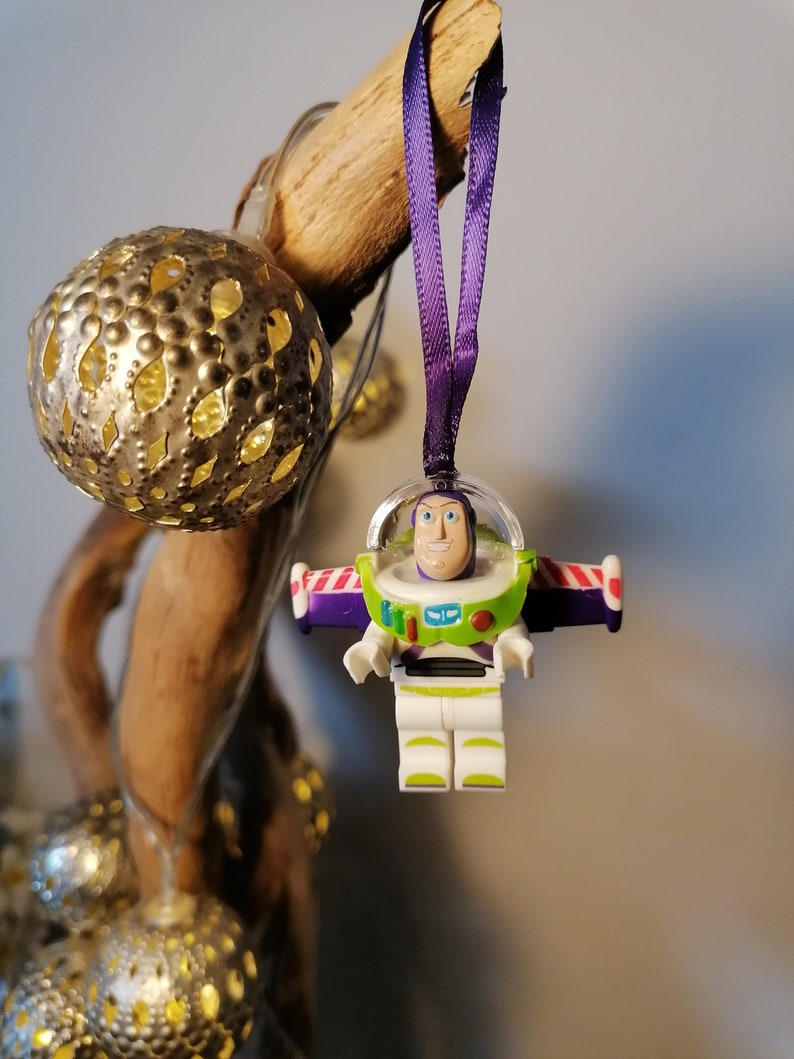 May include: A purple and white Christmas bauble featuring a plastic figurine of Buzz Lightyear from the Toy Story franchise. The bauble is hanging from a purple ribbon.