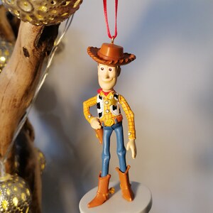 Op de afbeelding: Een rode koord-versiering met een plastic figuur van Woody uit Toy Story. Woody draagt zijn kenmerkende cowboyhoed, vest en jeans. Hij houdt zijn pistool in zijn rechterhand en heeft een vriendelijke uitdrukking op zijn gezicht.
