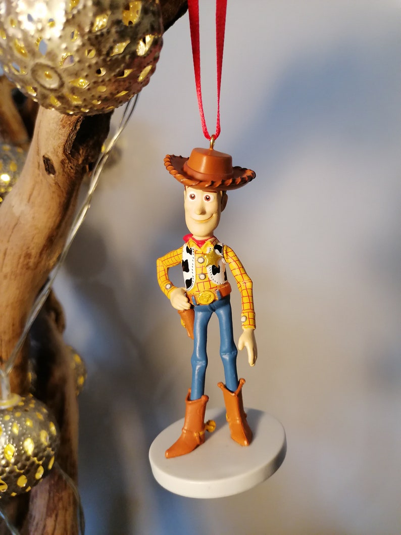 Op de afbeelding: Een rode koord-versiering met een plastic figuur van Woody uit Toy Story. Woody draagt zijn kenmerkende cowboyhoed, vest en jeans. Hij houdt zijn pistool in zijn rechterhand en heeft een vriendelijke uitdrukking op zijn gezicht.