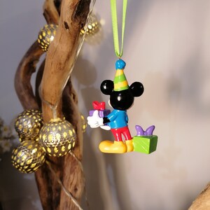 Peut inclure: Une figurine color&eacute;e de Mickey Mouse portant un chapeau de f&ecirc;te et tenant une bo&icirc;te cadeau. La figurine est suspendue &agrave; un ruban vert.