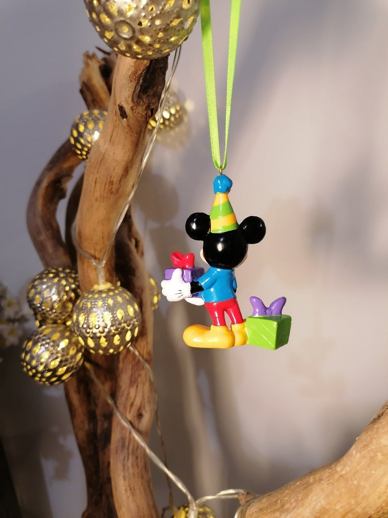 Peut inclure: Une figurine color&eacute;e de Mickey Mouse portant un chapeau de f&ecirc;te et tenant une bo&icirc;te cadeau. La figurine est suspendue &agrave; un ruban vert.