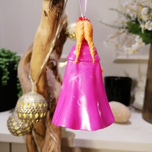 May include: A pink and white ornament of a girl with long braided hair wearing a pink cape. The ornament is hanging from a purple ribbon.