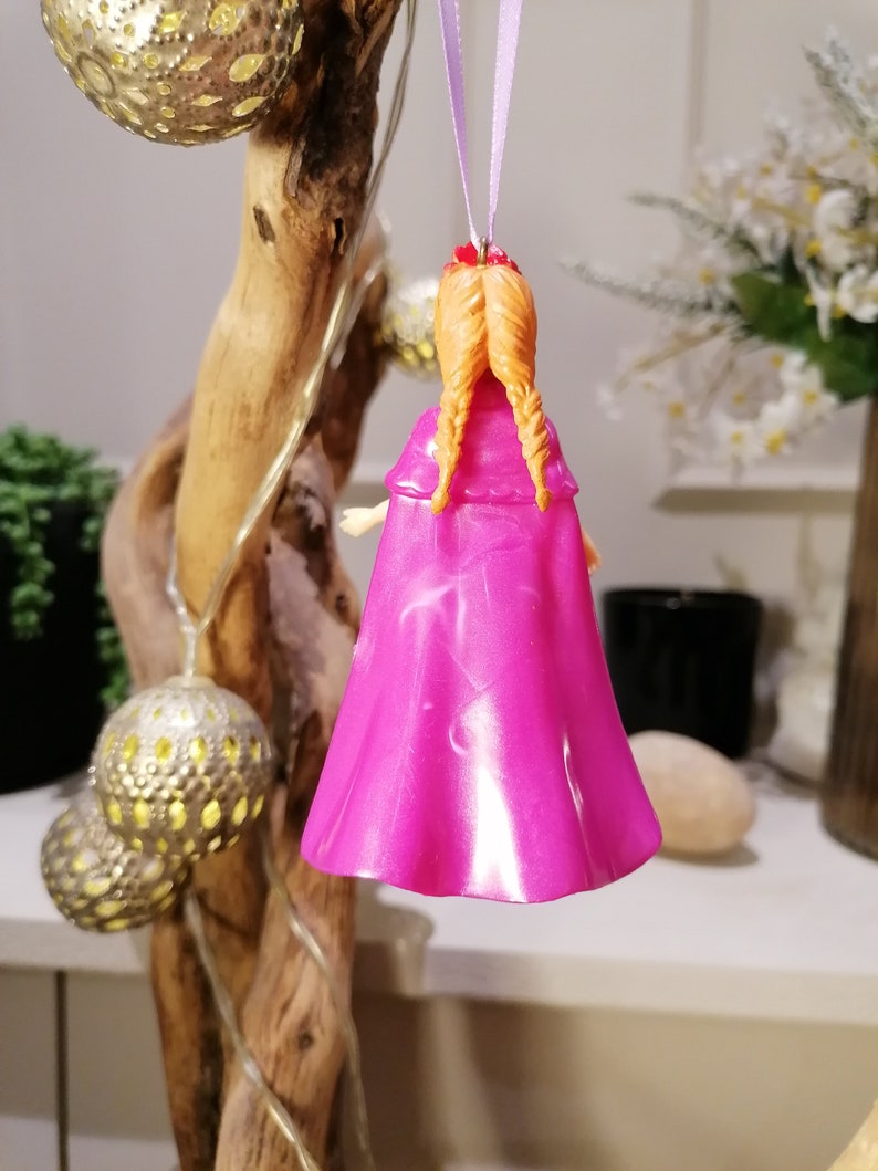 May include: A pink and white ornament of a girl with long braided hair wearing a pink cape. The ornament is hanging from a purple ribbon.