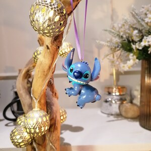 Pode incluir: Um ornamento azul do Stitch pendurado em uma fita roxa em um galho de madeira &agrave; deriva com luzes de corda e enfeites dourados.