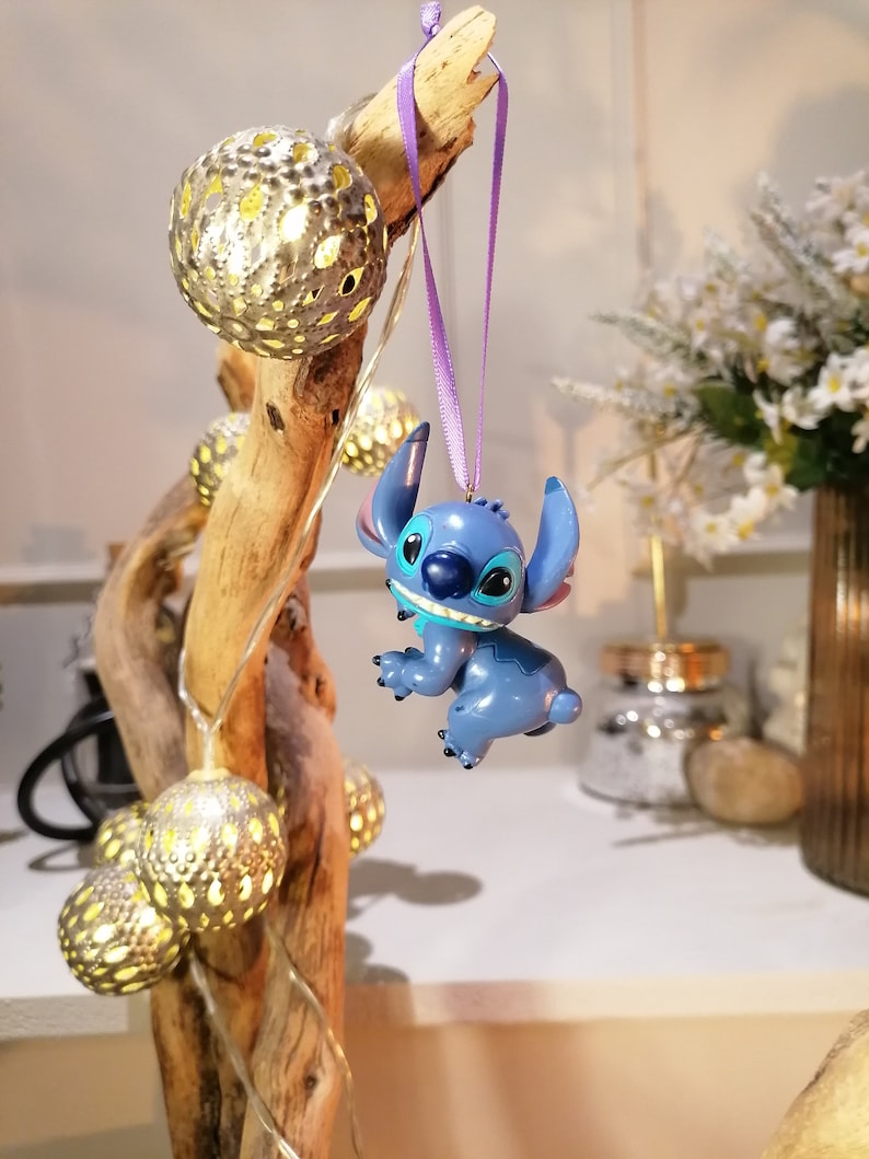 Pode incluir: Um ornamento azul do Stitch pendurado em uma fita roxa em um galho de madeira &agrave; deriva com luzes de corda e enfeites dourados.