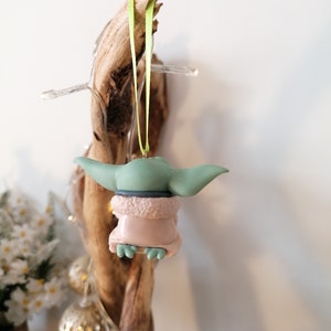 May include: A green and pink Christmas ornament featuring a character from the Star Wars franchise. The ornament is hanging from a green ribbon.