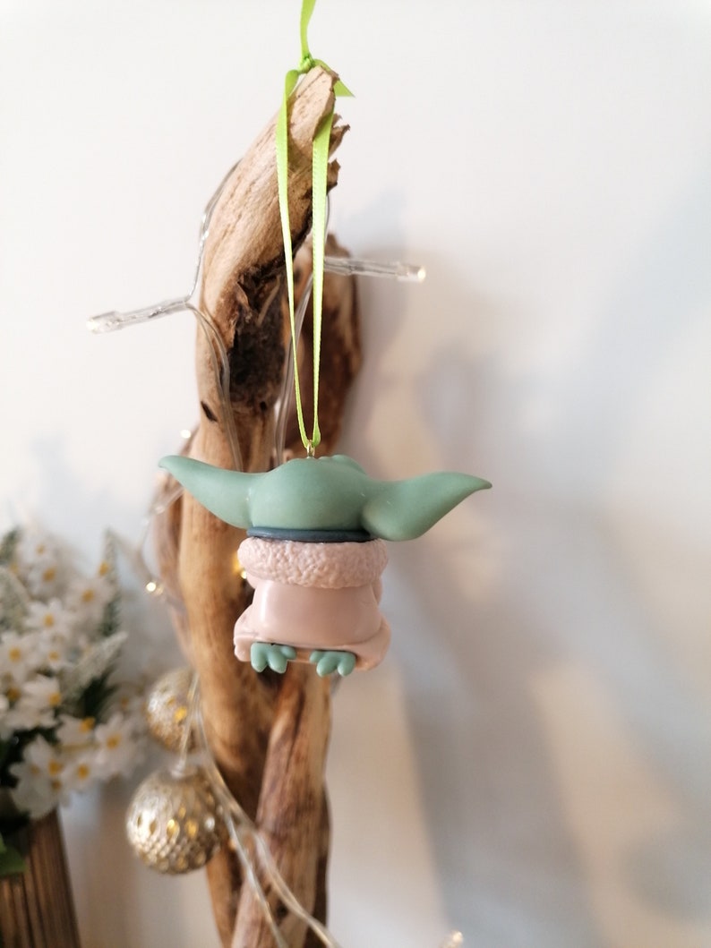 May include: A green and pink Christmas ornament featuring a character from the Star Wars franchise. The ornament is hanging from a green ribbon.