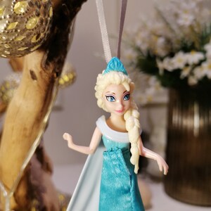May include: A blue and white plastic doll with blonde hair wearing a blue dress and a white cape. The doll is hanging from a white string.