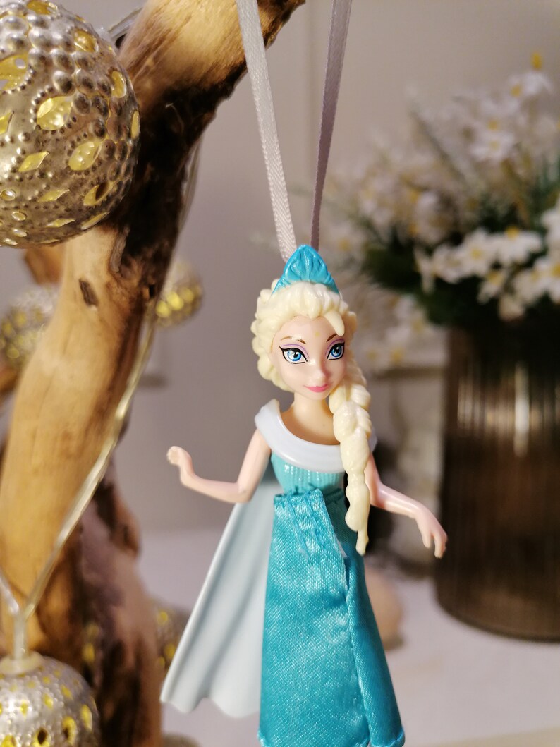 May include: A blue and white plastic doll with blonde hair wearing a blue dress and a white cape. The doll is hanging from a white string.