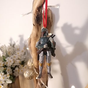 May include: A green and gray Star Wars Boba Fett Christmas ornament with a red ribbon hanging from a piece of driftwood.
