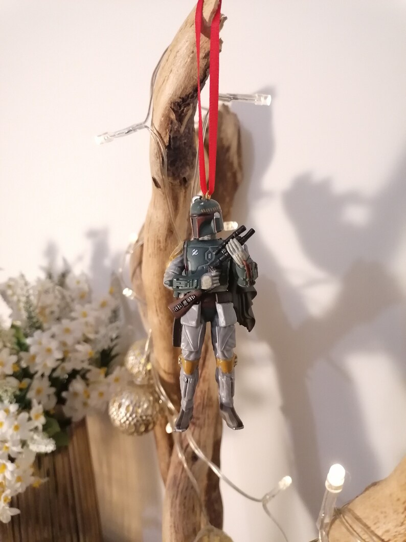 May include: A green and gray Star Wars Boba Fett Christmas ornament with a red ribbon hanging from a piece of driftwood.