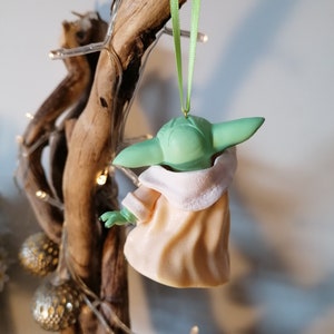 May include: A green and peach-colored Christmas ornament shaped like a character from the Star Wars franchise. The ornament is hanging from a green ribbon.