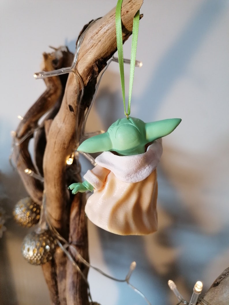 May include: A green and peach-colored Christmas ornament shaped like a character from the Star Wars franchise. The ornament is hanging from a green ribbon.