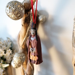 May include: A brown and red Christmas ornament depicting a figure wearing a crown and a long robe. The ornament is hanging from a red ribbon on a branch.