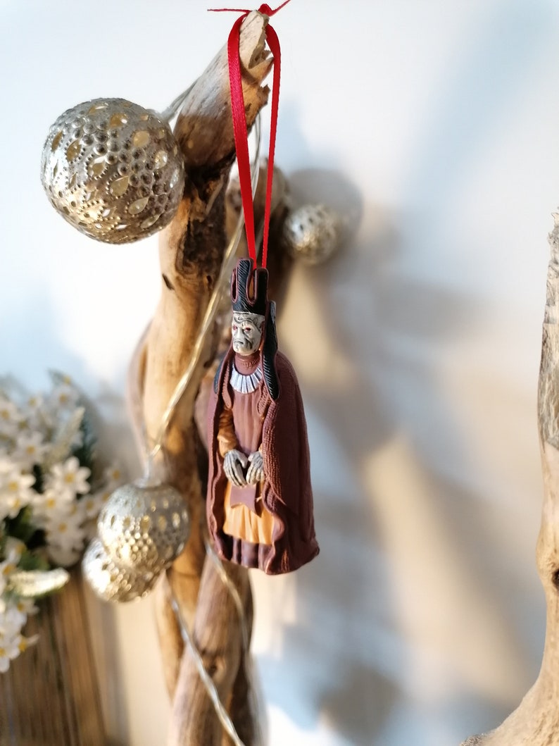 May include: A brown and red Christmas ornament depicting a figure wearing a crown and a long robe. The ornament is hanging from a red ribbon on a branch.