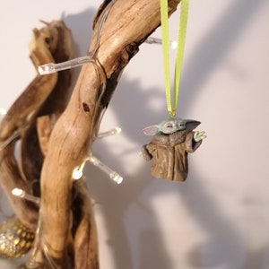 Pu&ograve; includere: Un ornamento natalizio verde e marrone con un personaggio del franchise di Star Wars. L'ornamento &egrave; appeso a un nastro verde su un ramo di legno.