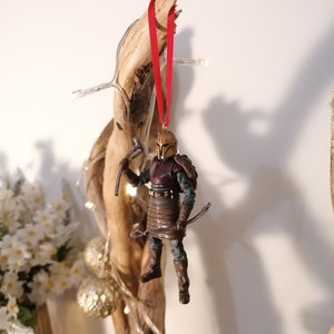 May include: A brown and gold action figure of a warrior wearing a helmet and holding a weapon. The figure is hanging from a red ribbon on a wooden branch.