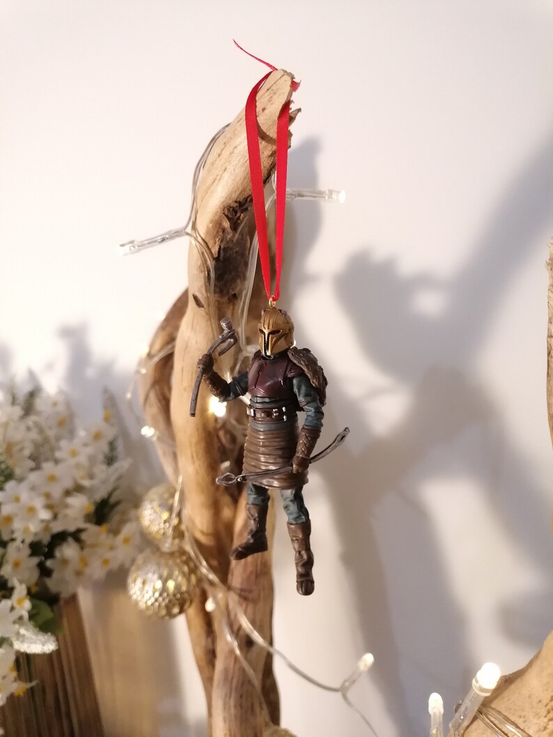 May include: A brown and gold action figure of a warrior wearing a helmet and holding a weapon. The figure is hanging from a red ribbon on a wooden branch.