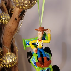 May include: A green toy car with a blue spoiler and a cartoon face.  Woody from Toy Story is sitting in the driver's seat.  The car is hanging from a green ribbon.