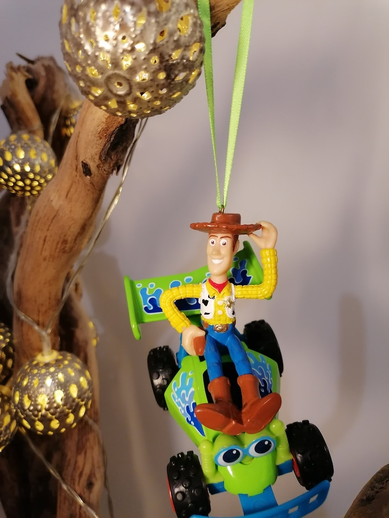 May include: A green toy car with a blue spoiler and a cartoon face.  Woody from Toy Story is sitting in the driver's seat.  The car is hanging from a green ribbon.