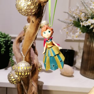 Pu&ograve; includere: Una piccola figurina di plastica di una ragazza con i capelli rossi che indossa un vestito blu e oro. La figurina &egrave; appesa a un ramo con luci dorate da uno spago verde.