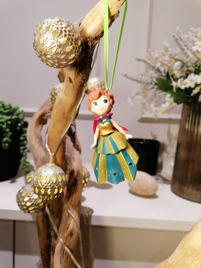 Pu&ograve; includere: Una piccola figurina di plastica di una ragazza con i capelli rossi che indossa un vestito blu e oro. La figurina &egrave; appesa a un ramo con luci dorate da uno spago verde.