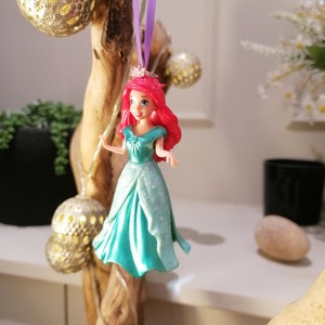 May include: A red-haired Disney princess doll wearing a teal gown with glitter accents. The doll is hanging from a purple ribbon and is a Christmas ornament.