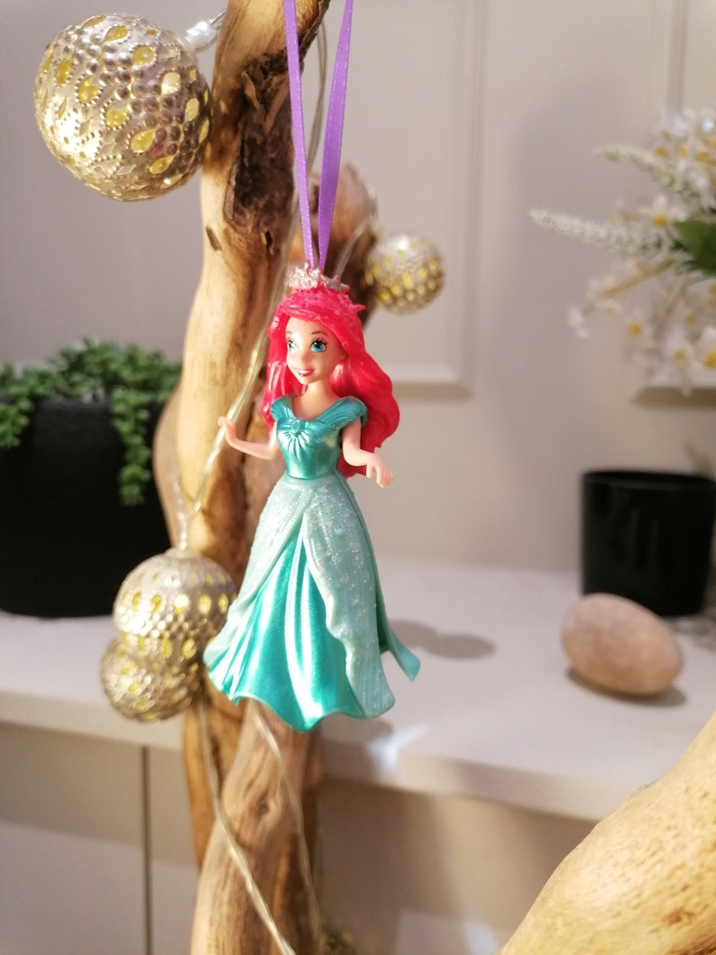 May include: A red-haired Disney princess doll wearing a teal gown with glitter accents. The doll is hanging from a purple ribbon and is a Christmas ornament.