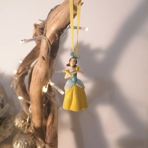 May include: A yellow and blue porcelain figurine of a woman wearing a yellow dress and a blue bodice. The figurine is hanging from a yellow ribbon on a branch.