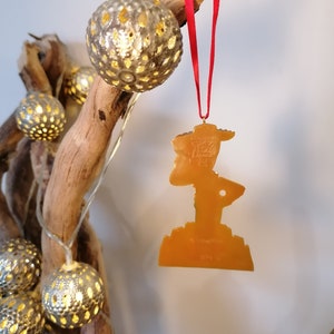 May include: A gold-colored plastic ornament shaped like Woody from the Toy Story franchise. The ornament is hanging from a red ribbon on a branch with gold-colored string lights.