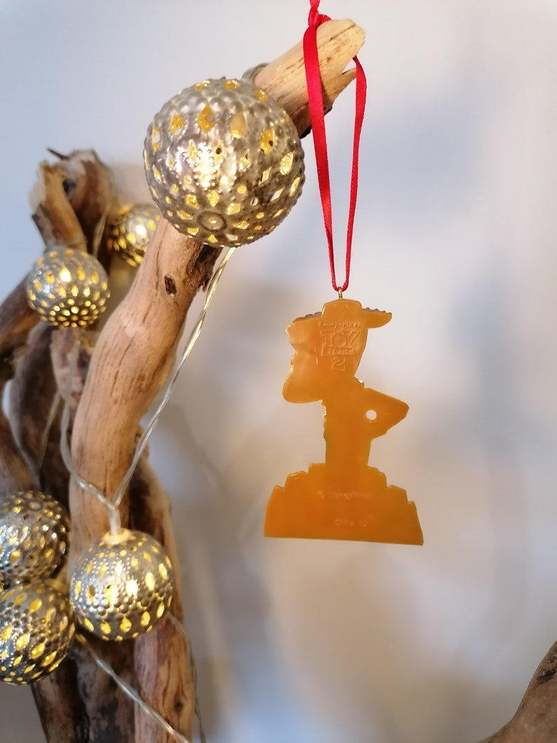 May include: A gold-colored plastic ornament shaped like Woody from the Toy Story franchise. The ornament is hanging from a red ribbon on a branch with gold-colored string lights.