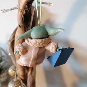 May include: A green and blue Christmas ornament featuring a character from the Star Wars franchise wearing a pink coat and holding a blue brick.