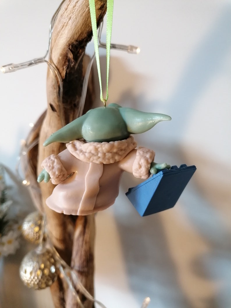 May include: A green and blue Christmas ornament featuring a character from the Star Wars franchise wearing a pink coat and holding a blue brick.