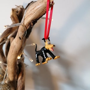 Pode incluir: Um enfeite de Natal de cão de desenho animado preto e amarelo com uma fita vermelha pendurada em um galho.