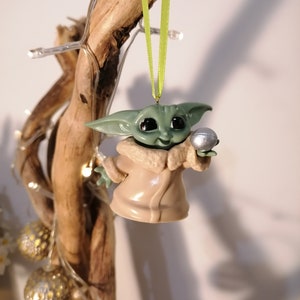 May include: A green and brown Christmas ornament featuring a character from the Star Wars franchise, holding a silver ball. The ornament is hanging from a green ribbon.