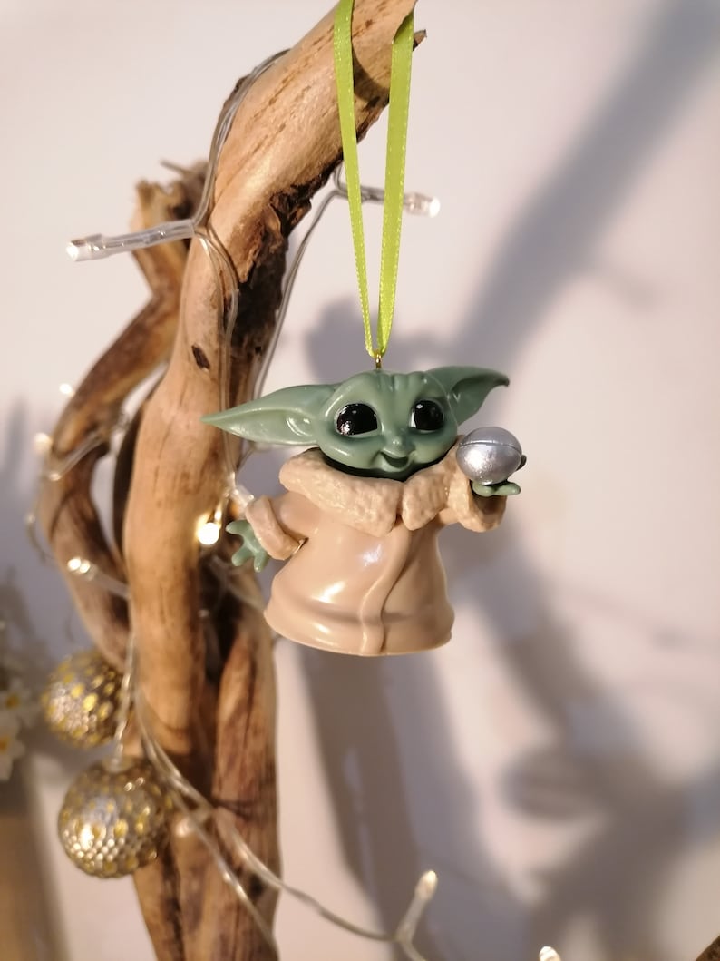 May include: A green and brown Christmas ornament featuring a character from the Star Wars franchise, holding a silver ball. The ornament is hanging from a green ribbon.
