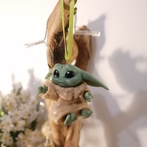 May include: A green and brown figurine of a character from the Star Wars franchise, hanging from a green ribbon on a piece of driftwood.