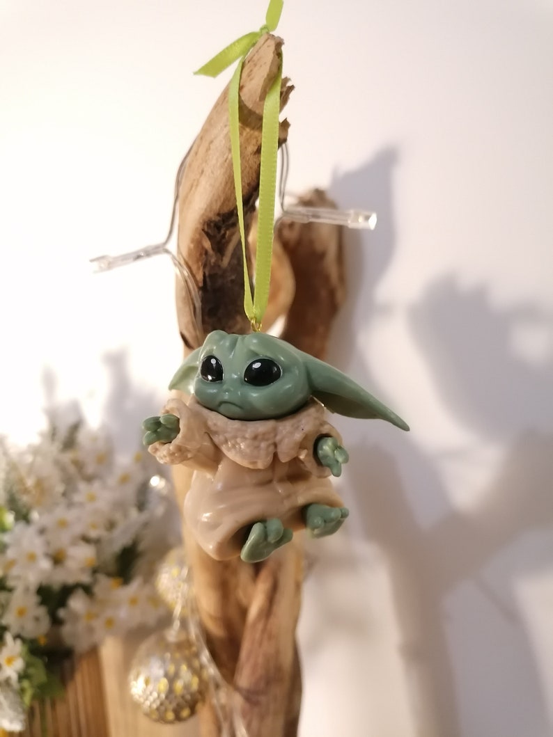 May include: A green and brown figurine of a character from the Star Wars franchise, hanging from a green ribbon on a piece of driftwood.