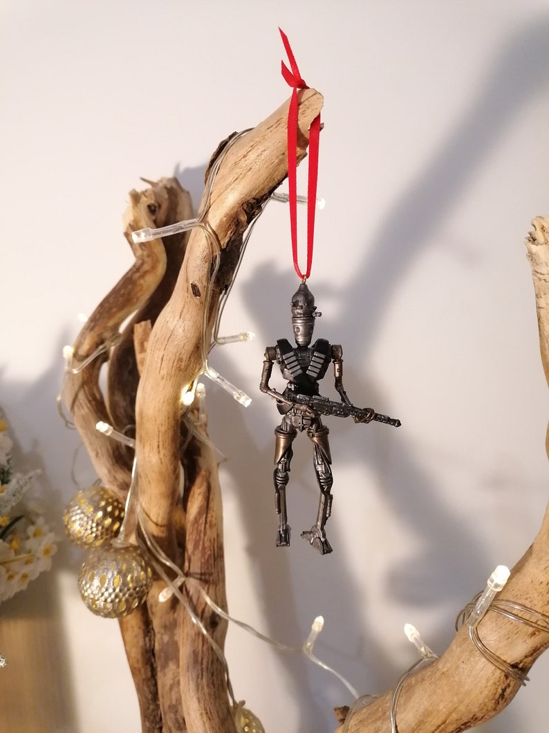 Puede incluir: Un adorno de robot met&aacute;lico con una pistola colgando de una rama con una cinta roja. El robot es un adorno navide&ntilde;o.