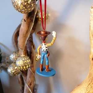 May include: A red string hangs from a wooden branch with a blue and white Christmas ornament of Woody from Toy Story. Woody is wearing his signature cowboy hat, vest, and jeans. He is waving with his right hand.