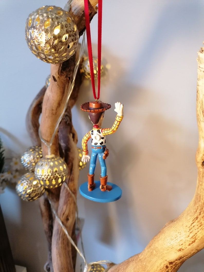 May include: A red string hangs from a wooden branch with a blue and white Christmas ornament of Woody from Toy Story. Woody is wearing his signature cowboy hat, vest, and jeans. He is waving with his right hand.