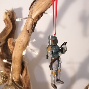 May include: A green and gray Star Wars Boba Fett Christmas ornament with a red ribbon hanging from a wooden branch.