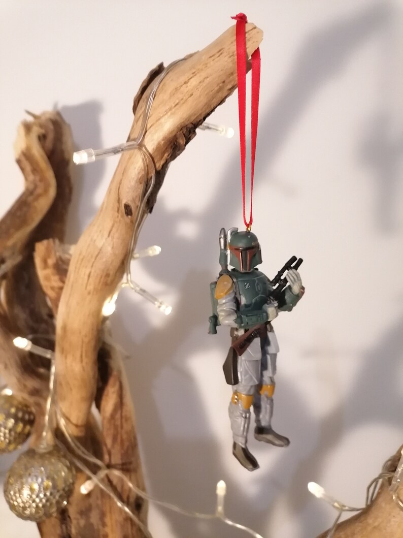 May include: A green and gray Star Wars Boba Fett Christmas ornament with a red ribbon hanging from a wooden branch.