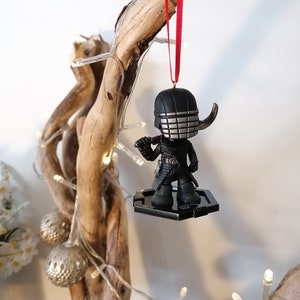 May include: A black and silver Christmas ornament featuring a character from the Star Wars franchise. The ornament is hanging from a red ribbon on a branch with white lights.