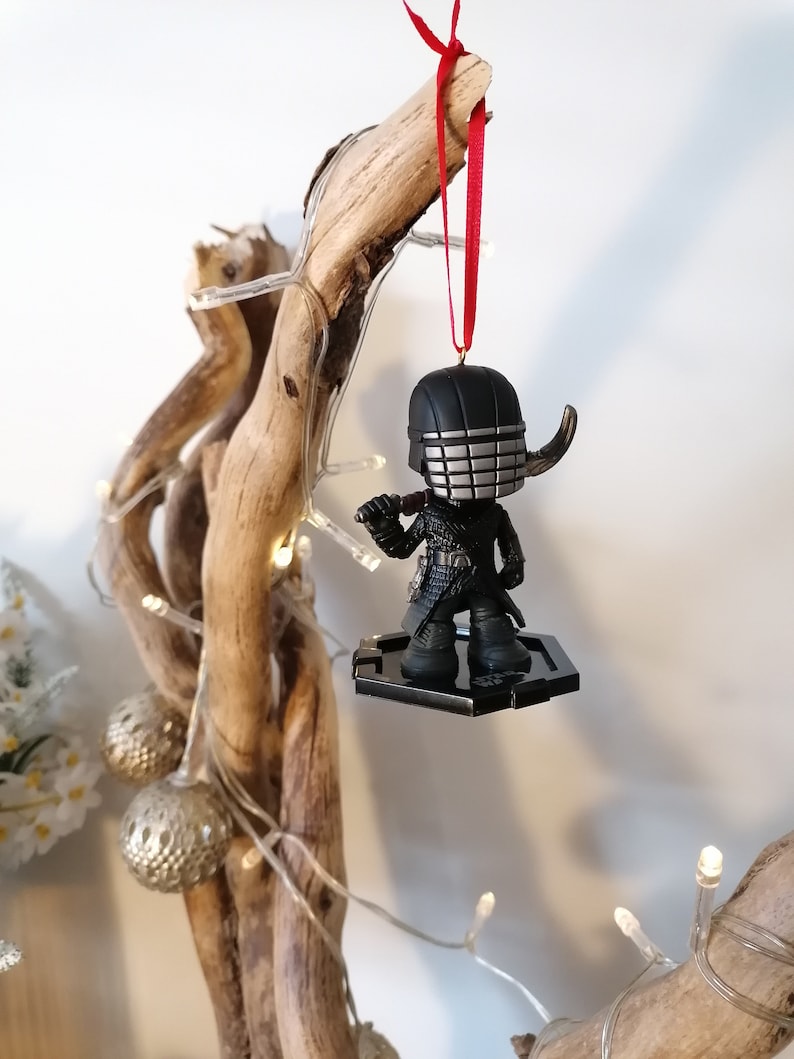 May include: A black and silver Christmas ornament featuring a character from the Star Wars franchise. The ornament is hanging from a red ribbon on a branch with white lights.