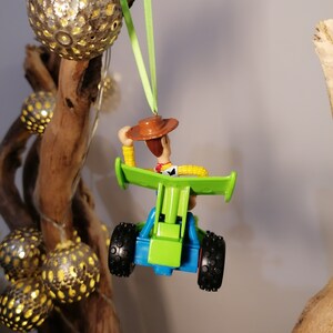 May include: A green toy car with a blue base and black wheels. A toy figure of Woody from Toy Story is sitting in the car. The car is hanging from a green ribbon.