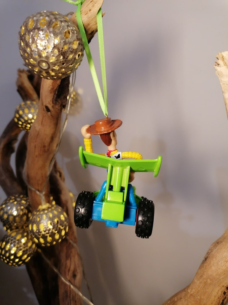 May include: A green toy car with a blue base and black wheels. A toy figure of Woody from Toy Story is sitting in the car. The car is hanging from a green ribbon.