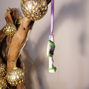 Puede incluir: Una cinta morada cuelga de una rama con una figurita de pl&aacute;stico blanca y verde de un ranger espacial. La figurita est&aacute; colgada boca abajo. La rama est&aacute; decorada con una cadena de luces con bolas doradas met&aacute;licas.