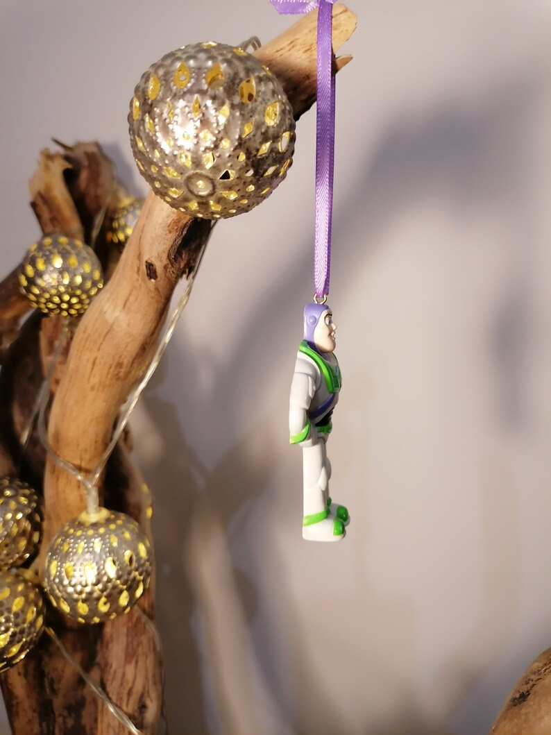 Puede incluir: Una cinta morada cuelga de una rama con una figurita de pl&aacute;stico blanca y verde de un ranger espacial. La figurita est&aacute; colgada boca abajo. La rama est&aacute; decorada con una cadena de luces con bolas doradas met&aacute;licas.