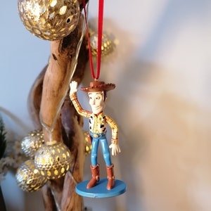 May include: A red string hangs from a wooden branch, holding a plastic figurine of Woody from Toy Story. Woody is wearing his signature cowboy hat, vest, and jeans. He is waving with his right hand and has a friendly expression on his face.